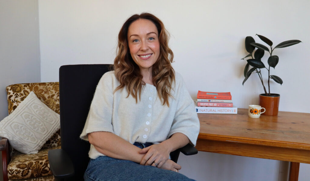 A portrait of Jules smiling and sitting in her office room. An armchair and small table with plants, books and a cup sits in the background.