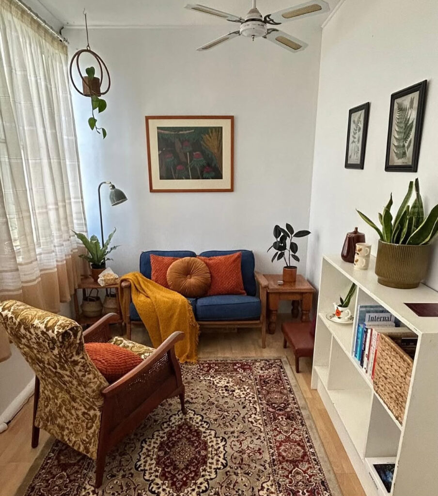 JC Counselling's stylish and comfortable office room with inviting blue couch, and floral armchair. Prints of flowers adorn the walls, a large rug spreads across the floor and several green plants are placed around the room.